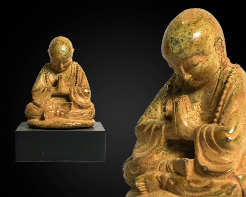 May include: A small, golden-brown ceramic statue of a meditating monk with hands clasped in prayer. The statue has a textured surface and is sitting on a black base.