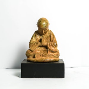 May include: A small, golden-brown ceramic statue of a meditating Buddha figure with hands clasped in prayer. The statue is sitting on a black wooden base.