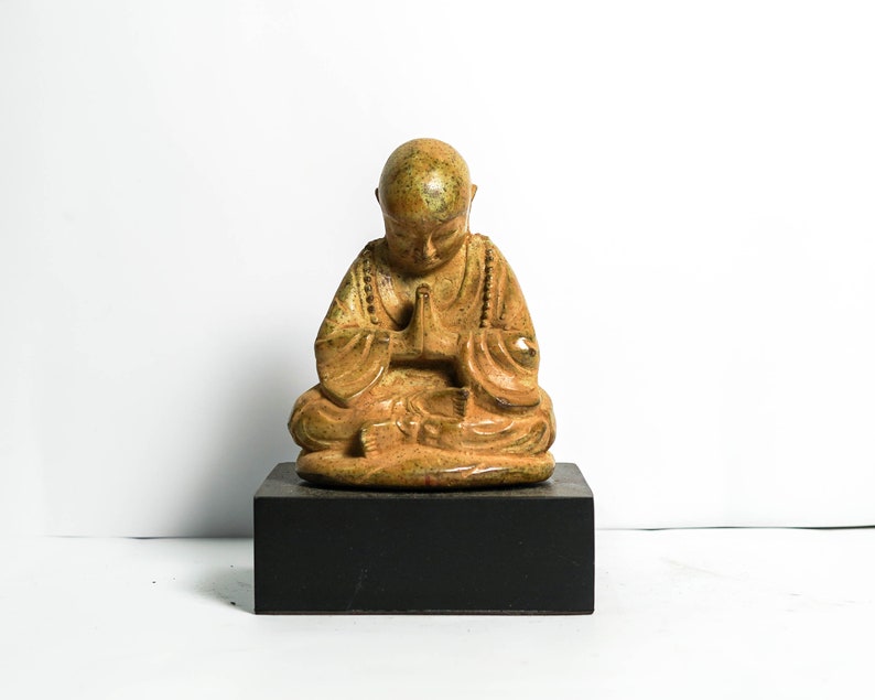 May include: A small, golden-brown ceramic statue of a meditating Buddha figure with hands clasped in prayer. The statue is sitting on a black wooden base.
