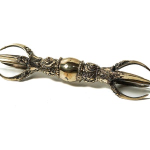 May include: A gold-colored metal ritual object with three prongs on each end. The object is decorated with intricate details and has a rounded center section.