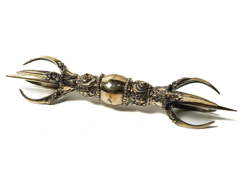 May include: A gold-colored metal ritual object with three prongs on each end. The object is decorated with intricate details and has a rounded center section.
