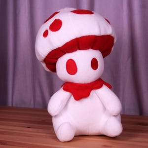 May include: White plush mushroom toy with red accents. The mushroom has a red cap with white spots and a red scarf around its neck.