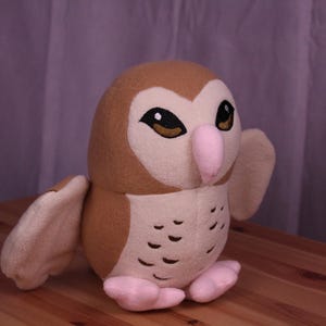 May include: A plush owl toy in shades of brown, cream, and pink. The owl has large, expressive eyes, a pink beak, and small feet. The wings are slightly outstretched, giving the impression of flight. The toy is sitting on a wooden surface.