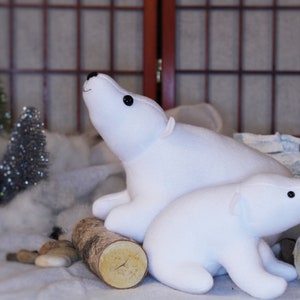 May include: Two white plush polar bear stuffed animals, one larger than the other, are sitting on a white surface with a wooden log behind them. The larger polar bear is looking up and the smaller polar bear is looking to the right.