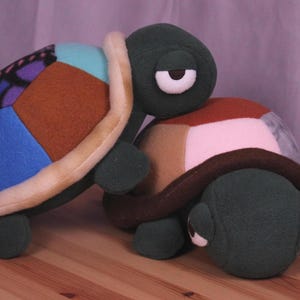 May include: Two plush turtle toys. One turtle has a shell made of colorful fabric patches in blue, brown, and purple. The other turtle has a shell made of brown, pink, and gray patches. Both turtles have green bodies and white eyes.