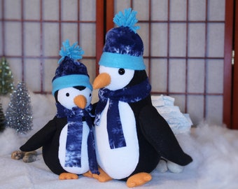 May include: Two plush penguin toys wearing blue and white hats and scarves. The penguins are standing on a white surface that looks like snow.