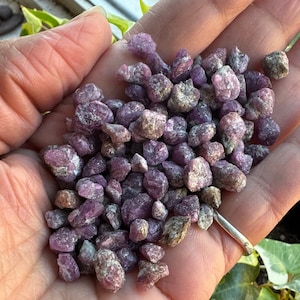 May include: A close-up of a hand holding a pile of small, purple and gray, rough-cut gemstones. The stones are irregularly shaped and have a slightly metallic sheen.