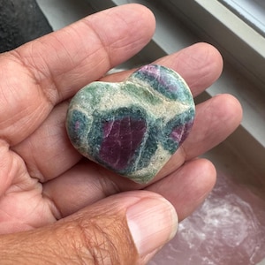 May include: Heart-shaped stone with a mix of green, white, and deep red hues. The stone has a polished surface and is held in a hand. The heart shape is clearly defined, showcasing the natural patterns and colors of the stone.