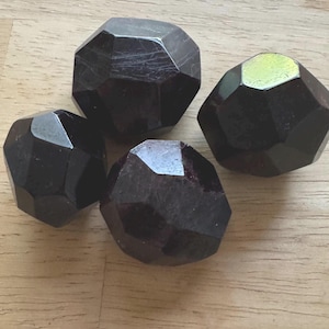 May include: Four faceted, dark-colored stones with a polished appearance. The stones are arranged on a light-colored wooden surface. The stones have a geometric shape and a deep, rich color.