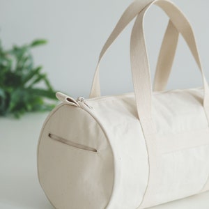 May include: A cream-colored canvas duffel bag with a cylindrical shape. The bag features a zippered top closure, a side pocket, and two sturdy handles. The bag is designed for carrying items and has a simple, minimalist aesthetic.