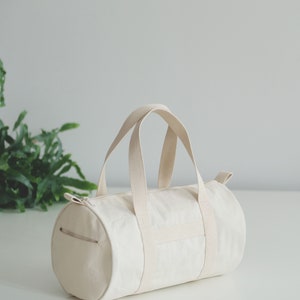 May include: Cream-colored canvas duffel bag with a cylindrical shape. The bag features two short handles and a long zipper closure. The bag is sitting on a white surface.