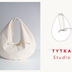 Pode incluir: Bolsa tote em forma de lágrima, cor creme, com fecho de cordão e um pequeno laço. A bolsa tem uma única alça e é feita de um tecido macio e texturizado. O texto "TYTKA Studio" está em vermelho.