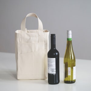 May include: A white canvas tote bag with a pocket on the front, holding a bottle of red wine and a bottle of white wine.