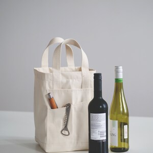 May include: A white canvas tote bag with a pocket on the front. The pocket contains a corkscrew and a wooden handled bottle opener. Two bottles of wine are in front of the bag, one red and one white.