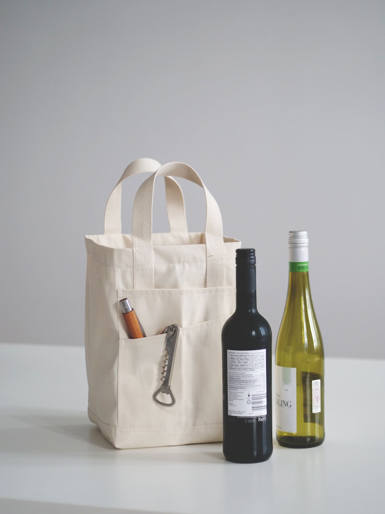 May include: A white canvas tote bag with a pocket on the front. The pocket contains a corkscrew and a wooden handled bottle opener. Two bottles of wine are in front of the bag, one red and one white.