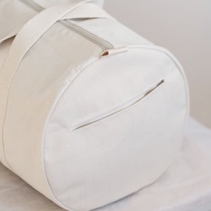 May include: Cream-colored canvas duffel bag with a cylindrical shape. Features a top zipper closure, a small exterior pocket, and two sturdy handles. The bag is made of a durable, textured fabric.