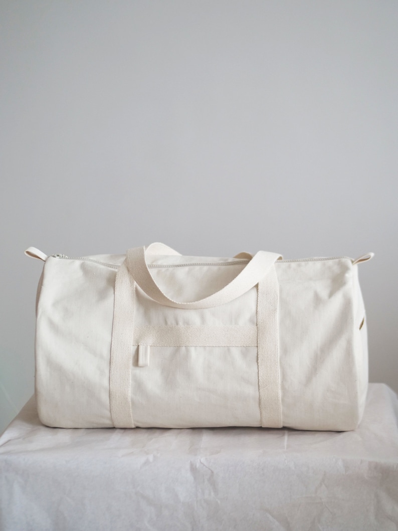 May include: A white canvas duffel bag with two top handles and a zipper closure. The bag has a single front pocket.