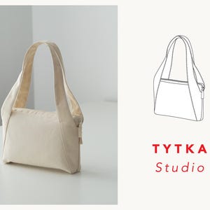 May include: A cream-colored canvas tote bag with a zipper closure and a simple design. The bag has a short handle and is photographed on a white surface. The text "TYTKA Studio" is printed in red below the bag.