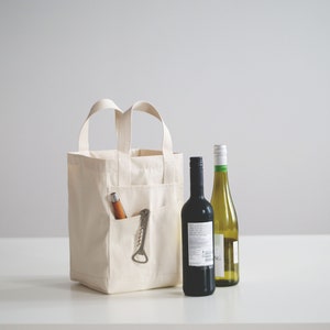 May include: A white canvas tote bag with a pocket on the side, containing a bottle opener and a wooden corkscrew. Two bottles of wine, one red and one white, are placed in front of the bag.