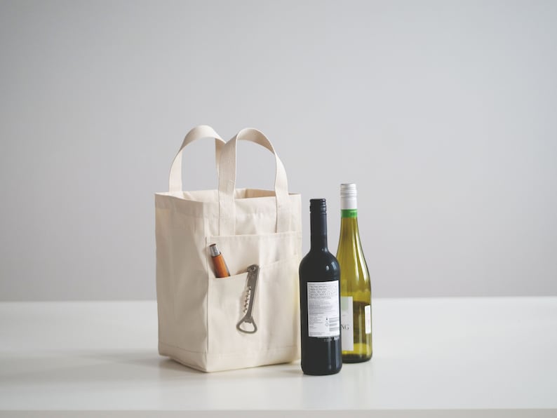 May include: A white canvas tote bag with a pocket on the side, containing a bottle opener and a wooden corkscrew. Two bottles of wine, one red and one white, are placed in front of the bag.