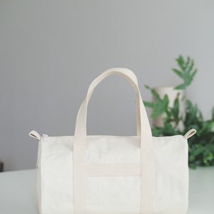 May include: Cream-colored canvas duffel bag with a cylindrical shape. Features two top handles and a shoulder strap. The bag has a zipper closure and a simple, minimalist design. Perfect for travel or everyday use.
