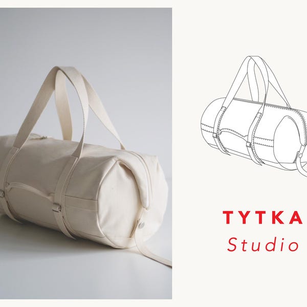 Op de afbeelding: Een witte canvas duffelbag met leren banden en gespen. De tas is gesloten en heeft een eenvoudig ontwerp. De tas staat op een wit oppervlak. De tekst "TYTKA Studio" staat in rode letters onder de tas.