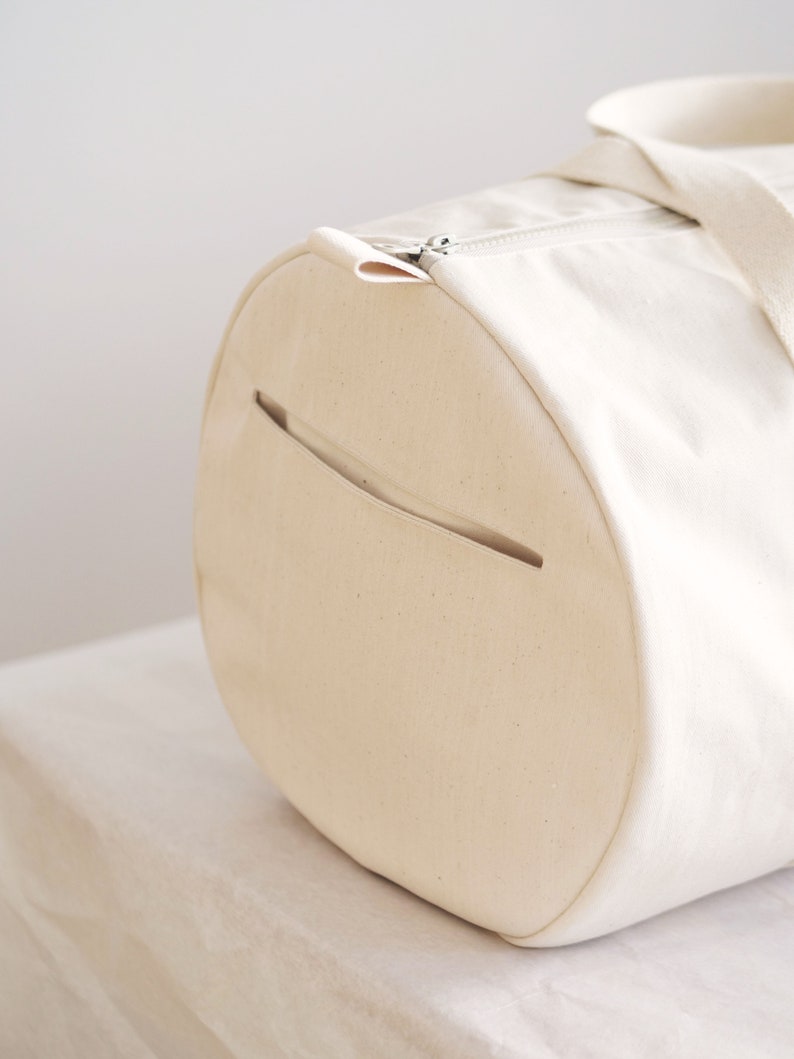 May include: Cream-colored canvas duffel bag with a cylindrical shape. The bag features a silver zipper closure, a side pocket, and a matching canvas handle. The bag is made of a sturdy material and is suitable for travel or everyday use.