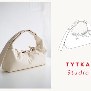 May include: A cream-coloured shoulder bag with a rectangular base and a curved handle. The bag features decorative bows and buttons. The TYTKA Studio logo is visible in the image.