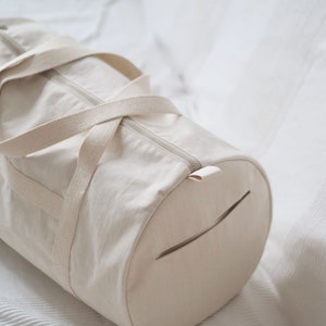 May include: A cream-colored canvas duffel bag with a zipper closure and a small pocket on the side. The bag has a simple design and is made of natural materials.