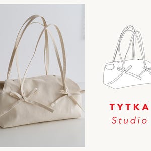 May include: A cream-coloured tote bag with long handles and bow details. The bag is rectangular with rounded edges and features a simple, minimalist design. The text "TYTKA Studio" is displayed in red.