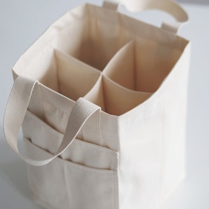 May include: A white canvas tote bag with six individual compartments and two handles. The bag is designed to carry and organize six bottles.