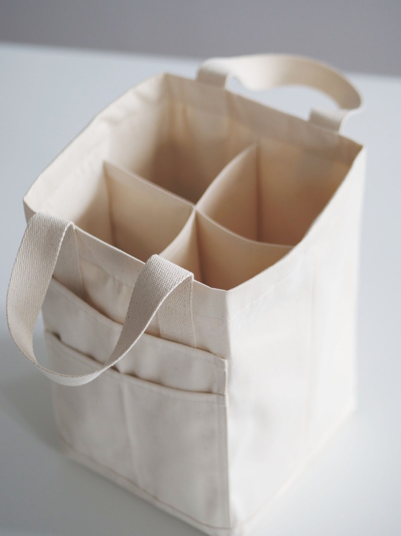 May include: A white canvas tote bag with six individual compartments and two handles. The bag is designed to carry and organize six bottles.