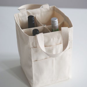 May include: A white canvas tote bag with two handles and a zippered pocket. The bag has six compartments for holding bottles. Two bottles are visible, one black and one green.