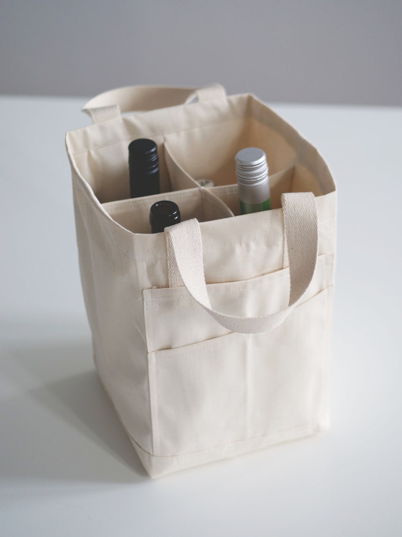 May include: A white canvas tote bag with two handles and a zippered pocket. The bag has six compartments for holding bottles. Two bottles are visible, one black and one green.