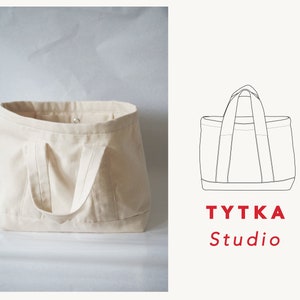 Può includere: Una borsa tote in tela bianca con una singola tasca sul davanti. La borsa ha due manici ed è realizzata in tessuto robusto. La borsa è appoggiata su una superficie bianca. Il testo "TYTKA Studio" è stampato in rosso sotto un disegno a linea della borsa.