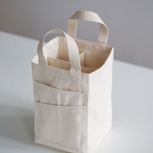 May include: A white canvas tote bag with two handles and multiple compartments. The bag is folded and sitting on a white surface.