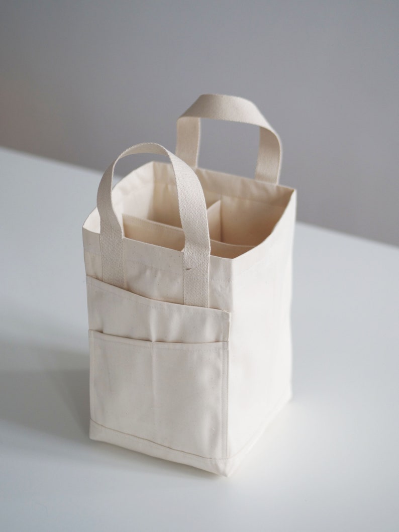 May include: A white canvas tote bag with two handles and multiple compartments. The bag is folded and sitting on a white surface.