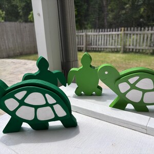 May include: Three wooden turtle figurines painted in shades of green and white. The turtles are arranged in a row on a white surface.