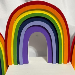 May include: Wooden rainbow arch toy in vibrant colors. The arch features layers of red, orange, yellow, green, blue, and purple. The toy is designed with a white base and is suitable for children's play or as a decorative item.
