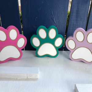 May include: Three wooden paw prints in pink, teal, and purple with white centers and gold outlines.