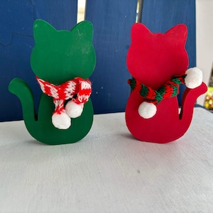 May include: Two wooden cat figurines, one green and one red, wearing festive scarves with white pom-poms. The cats are standing on a white surface.