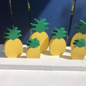 May include: A collection of wooden pineapple decorations in various sizes. Each pineapple is painted yellow with green leafy tops. The pineapples are arranged on a white surface against a blue background.