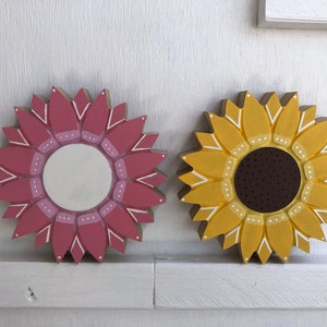 May include: Two wooden sunflower decorations, one painted pink with a white center and the other painted yellow with a brown center.