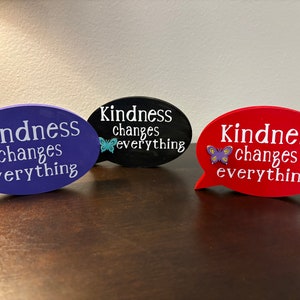May include: Three wooden speech bubble signs with the text "Kindness changes everything" in white lettering. Each sign has a different color: purple, black, and red. Each sign has a small butterfly decoration.