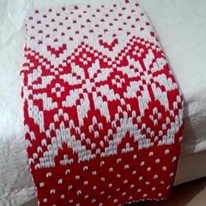 May include: A rectangular crochet blanket featuring a bold red and white geometric pattern. The design includes a large central snowflake motif surrounded by smaller repeating dots and chevron-like shapes, creating a high-contrast textile piece.