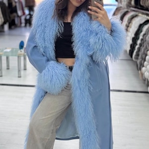 May include: A light blue long coat featuring a prominent, shaggy, textured trim along the lapels and cuffs. The coat is worn over a black cropped top and light gray denim jeans, paired with black and white sneakers.