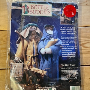 May include: A craft kit for making a three-piece set of bottle buddies depicting the Holy Family. The kit includes yarn, fabric, embroidery thread, ribbon floss, needles, and instructions. The figures are approximately 12 inches tall.