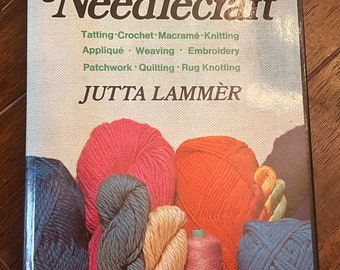 Könnte beinhalten: Ein Buch mit dem Titel "The Reinhold Book of Needlecraft" mit einem Einband, der farbige Wollknäuel zeigt. Das Buch behandelt Techniken wie Occhi, Häkeln und Sticken. Der Name des Autors, Jutta Lammer, ist ebenfalls sichtbar.