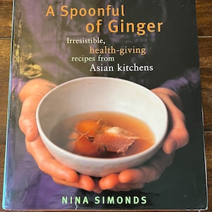 May include: A cookbook titled "A Spoonful of Ginger: Irresistible, health-giving recipes from Asian kitchens" by Nina Simonds. The cover features a close-up of hands holding a white bowl of ginger soup.