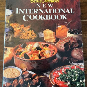 May include: A red, white, and blue cookbook titled "Betty Crocker's New International Cookbook". The cover features a variety of international dishes, including paella, a salad with tomatoes and feta cheese, and skewers of meat.